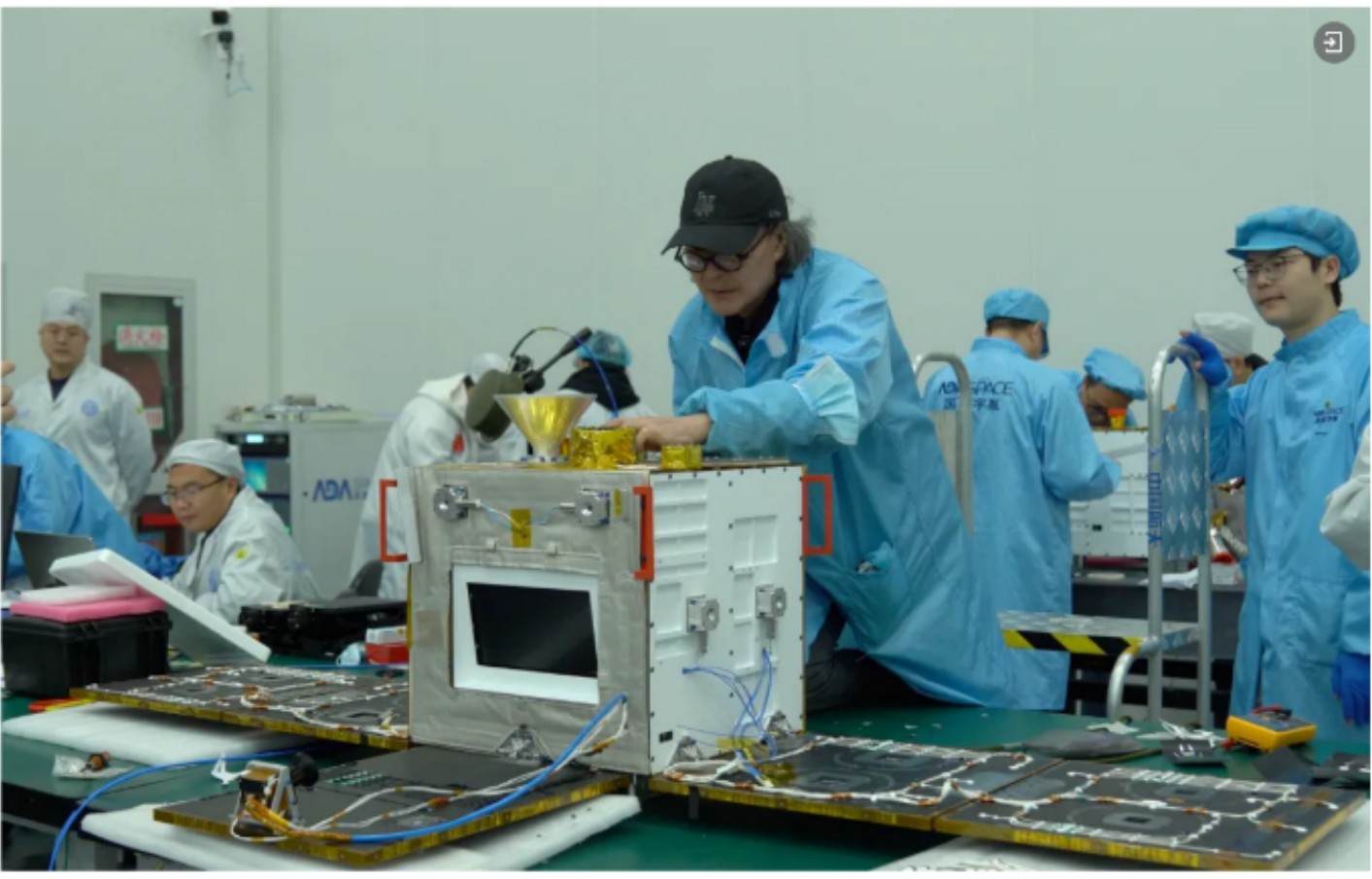 Technicians in blue clean-room suits working on the satellite during final assembly, its solar panels laid flat on the bench.